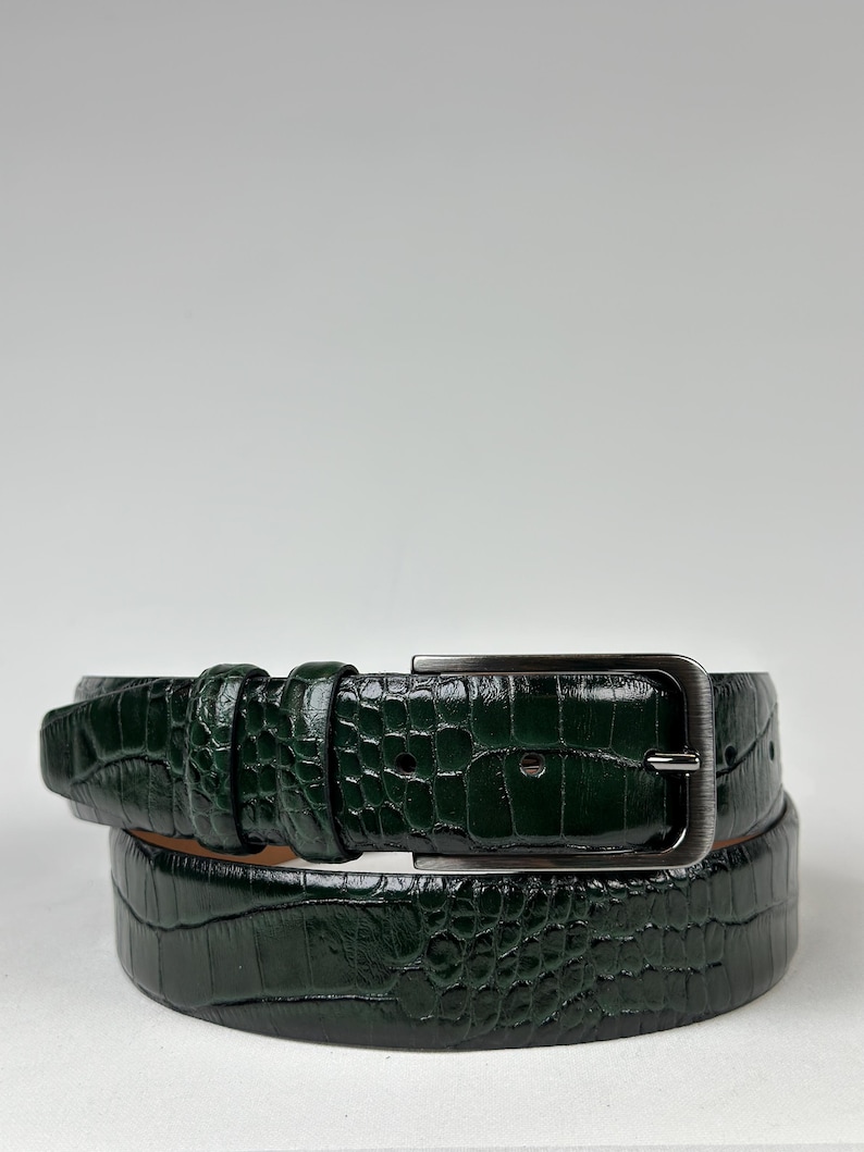 Peut inclure: Une ceinture vert fonc&eacute; avec un motif textur&eacute; en forme de crocodile. La ceinture est dot&eacute;e d'une boucle rectangulaire de couleur argent&eacute;e et d'une boucle assortie. La ceinture en cuir est enroul&eacute;e, montrant la texture et la couleur.