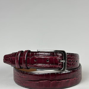 Peut inclure: Une ceinture en cuir bordeaux avec une texture peau de crocodile. La ceinture a une boucle rectangulaire de couleur argent&eacute;e et une s&eacute;rie de passants. La ceinture est enroul&eacute;e, montrant la texture et la couleur.
