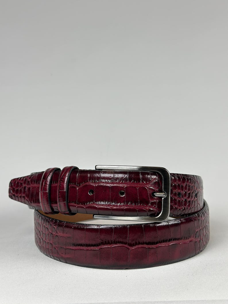 Peut inclure: Une ceinture en cuir bordeaux avec une texture peau de crocodile. La ceinture a une boucle rectangulaire de couleur argent&eacute;e et une s&eacute;rie de passants. La ceinture est enroul&eacute;e, montrant la texture et la couleur.