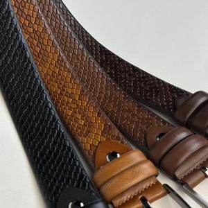 Puede incluir: Primer plano de varios cinturones de cuero en varios tonos de marrón y negro. Los cinturones tienen un patrón texturizado, similar a la piel de reptil, y hebillas plateadas. Los colores van desde el negro intenso hasta el beige claro.