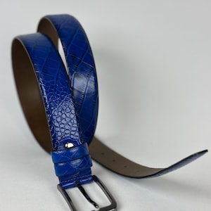 Peut inclure: Une ceinture bleue avec un motif textur&eacute; en forme de peau de crocodile. La ceinture a une boucle de couleur argent&eacute;e et un dessous marron. La ceinture est enroul&eacute;e, montrant toute sa longueur. Le fond est blanc.