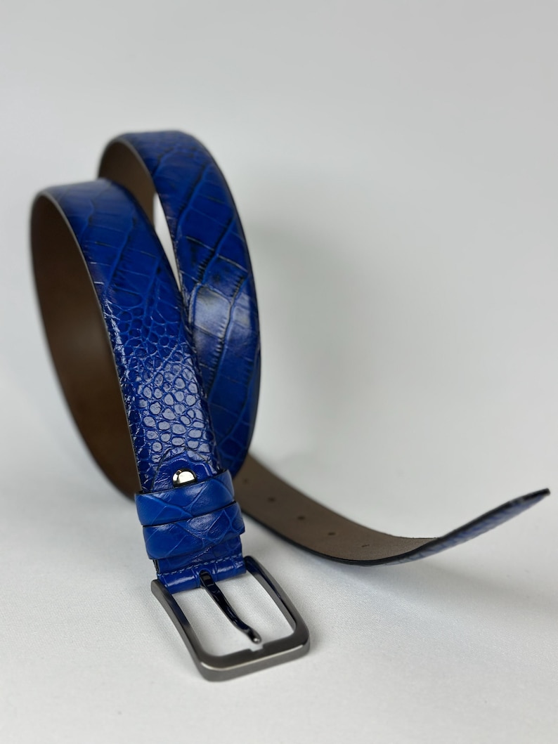 Peut inclure: Une ceinture bleue avec un motif textur&eacute; en forme de peau de crocodile. La ceinture a une boucle de couleur argent&eacute;e et un dessous marron. La ceinture est enroul&eacute;e, montrant toute sa longueur. Le fond est blanc.