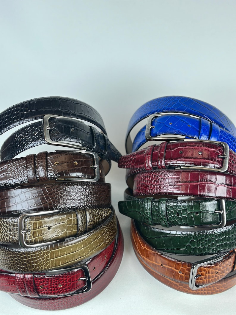Peut inclure: Plusieurs ceintures en cuir de diff&eacute;rentes couleurs, dont noir, marron, bleu, bordeaux, vert et beige. Chaque ceinture est orn&eacute;e d'une boucle argent&eacute;e et d'un motif textur&eacute; en forme de crocodile. Les ceintures sont empil&eacute;es en deux piles distinctes sur fond blanc.