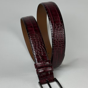 Peut inclure: Une ceinture en cuir bordeaux avec un motif textur&eacute; en forme de crocodile. La ceinture est dot&eacute;e d'une boucle rectangulaire de couleur canon de fusil et d'un int&eacute;rieur marron clair. La ceinture est enroul&eacute;e, mettant en valeur son design et sa texture.