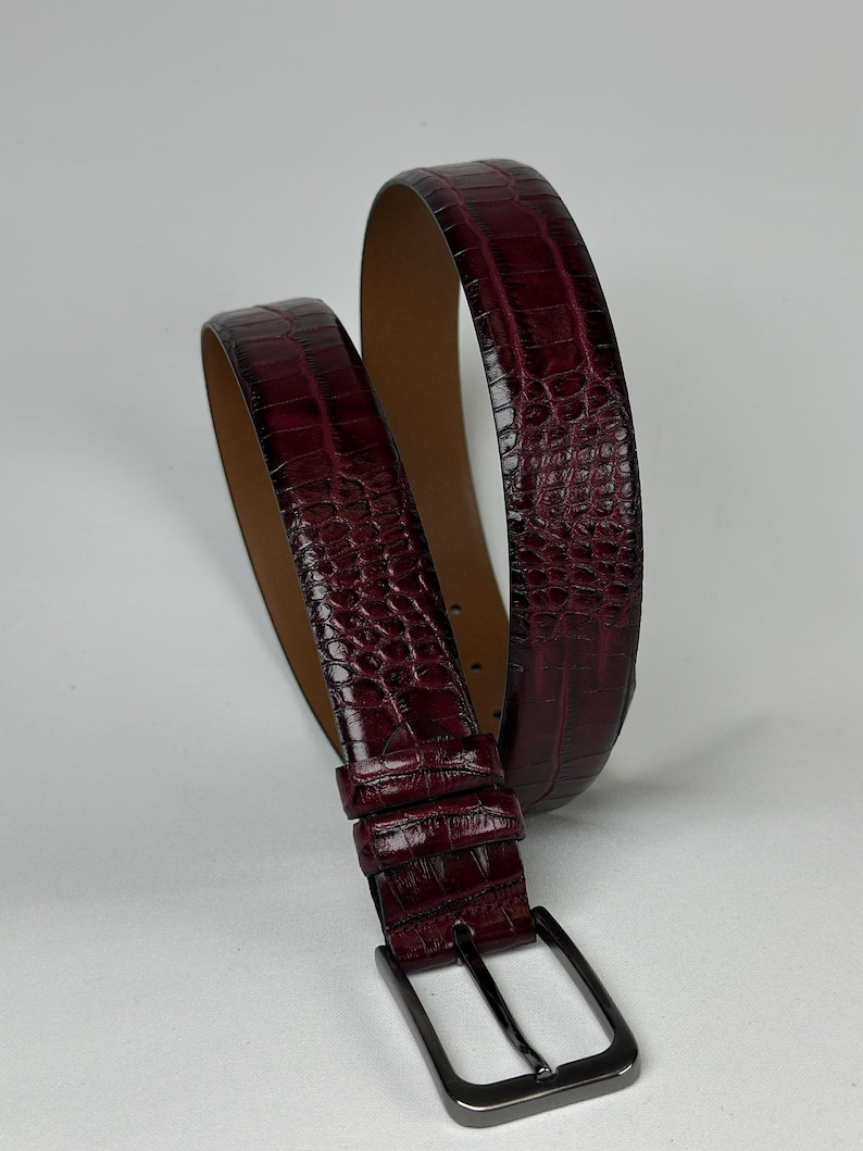 Peut inclure: Une ceinture en cuir bordeaux avec un motif textur&eacute; en forme de crocodile. La ceinture est dot&eacute;e d'une boucle rectangulaire de couleur canon de fusil et d'un int&eacute;rieur marron clair. La ceinture est enroul&eacute;e, mettant en valeur son design et sa texture.