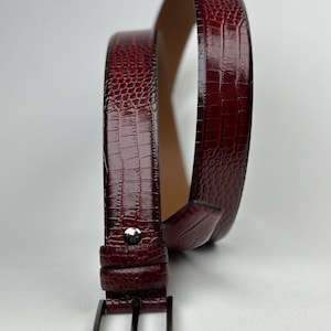 Peut inclure: Une ceinture bordeaux avec un motif textur&eacute; en forme de peau de crocodile. La boucle est de couleur argent&eacute;e, ainsi qu'un seul clou. La ceinture est enroul&eacute;e, r&eacute;v&eacute;lant l'int&eacute;rieur en cuir beige.