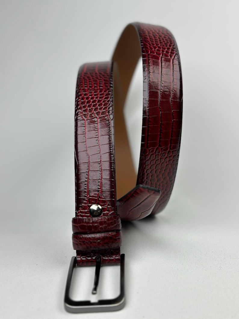 Peut inclure: Une ceinture bordeaux avec un motif textur&eacute; en forme de peau de crocodile. La boucle est de couleur argent&eacute;e, ainsi qu'un seul clou. La ceinture est enroul&eacute;e, r&eacute;v&eacute;lant l'int&eacute;rieur en cuir beige.