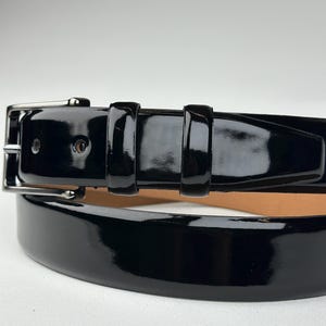 Pode incluir: Um cinto de couro preto com acabamento brilhante e laqueado. O cinto tem uma fivela prateada e est&aacute; dobrado ao meio.