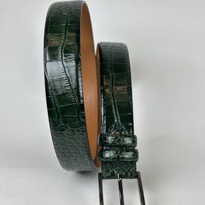 Peut inclure: Une ceinture en cuir vert fonc&eacute; avec une texture peau de crocodile. La ceinture a une boucle de couleur argent et un int&eacute;rieur marron clair. La ceinture est enroul&eacute;e, mettant en valeur sa texture et son design.