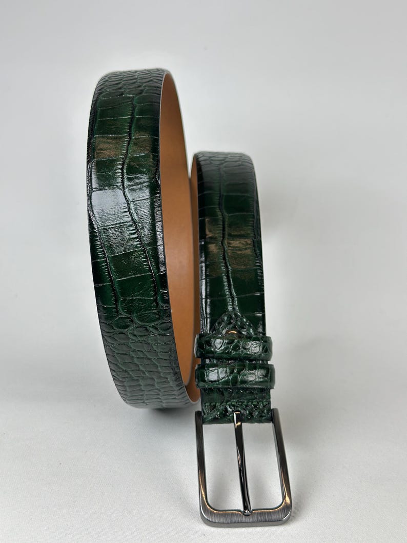 Peut inclure: Une ceinture en cuir vert fonc&eacute; avec une texture peau de crocodile. La ceinture a une boucle de couleur argent et un int&eacute;rieur marron clair. La ceinture est enroul&eacute;e, mettant en valeur sa texture et son design.