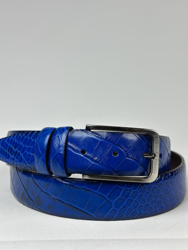 Peut inclure: Une ceinture bleu vif enroul&eacute;e, avec un motif textur&eacute; en forme de peau de crocodile. La ceinture est dot&eacute;e d'une boucle rectangulaire de couleur argent&eacute;e. Cette ceinture en cuir est un accessoire &eacute;l&eacute;gant.