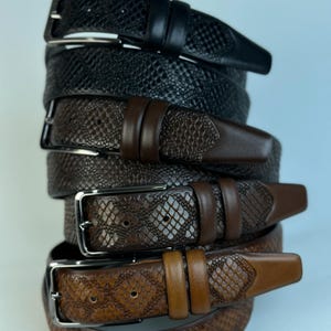 May include: A stack of leather belts in various shades of brown and black. The belts have silver buckles and textured patterns, including a snakeskin design. The belts are stacked on top of each other.