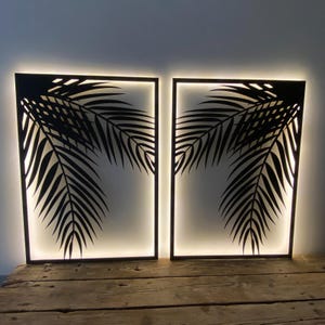 Puede incluir: Dos paneles de decoración de pared de metal negro con un diseño de hoja de palma. Los paneles están iluminados desde atrás con luces LED blancas cálidas.