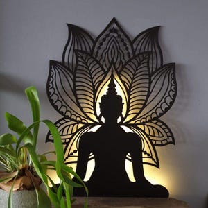 Puede incluir: Silueta negra de una figura de Buda meditando sentada dentro de un diseño de mandala detallado. El mandala está hecho de intrincados patrones florales y el Buda está sentado en posición de loto. El diseño está iluminado desde atrás, creando un brillo cálido.