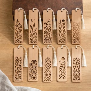 Könnte beinhalten: Zehn rechteckige Lesezeichen aus hellem Holz, angeordnet in zwei Reihen. Jedes Lesezeichen weist komplizierte lasergeschnittene Muster auf, darunter florale, himmlische und geometrische Designs. Jedes Lesezeichen ist mit einer kleinen weißen Stoffquaste versehen.