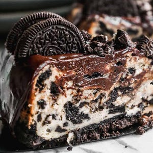 Puede incluir: Una porción de pastel de queso cubierto de chocolate con galletas Oreo, cubierto con migas de Oreo trituradas y una galleta Oreo entera. El pastel de queso está sobre una superficie de mármol blanco.