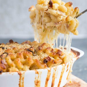 Puede incluir: Un plato de macarrones con queso horneados en una fuente de cerámica blanca, sobre una tabla de madera. Los macarrones están cubiertos con una corteza dorada. Un tenedor levanta una porción de pasta con queso, con hilos de queso fundido.