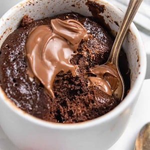 May include: A white mug filled with a chocolate mug cake topped with chocolate spread. A spoon is in the mug, and a bite has been taken out of the cake.