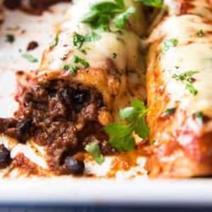 May include: Close-up of a baked casserole dish with enchiladas. The enchiladas are filled with a dark, savory mixture and topped with melted cheese and fresh parsley. The dish is in a white rectangular baking pan.