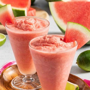 May include: Two tall glasses filled with a pink, blended watermelon drink, garnished with watermelon slices. Slices of watermelon and lime are in the background, suggesting a refreshing summer beverage.