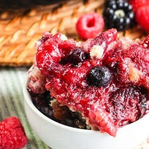 Puede incluir: Un bol blanco lleno de un crumble de bayas rojas y azules. El crumble está cubierto con una salsa roja dulce y se ven algunas arándanos. El bol está sobre una superficie rayada azul y blanca.