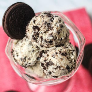 May include: A clear glass bowl filled with scoops of cookies and cream ice cream. A chocolate sandwich cookie is propped on the side. The ice cream has a creamy white base with dark chocolate cookie pieces throughout.