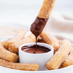 Puede incluir: Un bol blanco lleno de salsa de chocolate para mojar y varios churros largos, delgados y fritos espolvoreados con azúcar y canela. Un churro se está mojando en la salsa de chocolate.