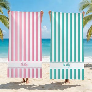 Puede incluir: Dos toallas de playa con rayas verticales. Una es rosa y blanca, la otra es turquesa y blanca. Cada toalla tiene el nombre "Lily" impreso en cursiva en una banda blanca. Las toallas se sostienen en una playa de arena con palmeras y el océano al fondo.