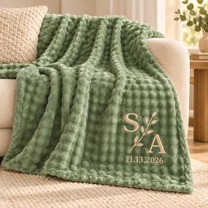 May include: A sage green textured waffle-knit throw blanket featuring embroidered beige lettering with the initials S and A separated by a leafy branch motif, with the date 11.13.2026 stitched in beige thread below the initials.