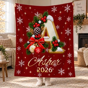 May include: A deep red fleece blanket featuring a large letter A decorated with pine branches, red berries, a pinecone, a red bow, and ornaments. The blanket displays the name Asher and the year 2026 in gold script, surrounded by white snowflakes.