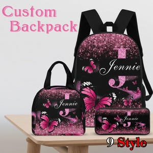 Könnte beinhalten: Ein individuelles Rucksack-Set mit Rucksack, Lunch-Tasche und Federmäppchen. Das Set ist schwarz mit einem rosa Glitzer-Design und Schmetterlingsakzenten. Der Name "Jennie" ist auf jedem Artikel zu sehen. Die Wörter "Custom Backpack" und "9 Style" sind ebenfalls sichtbar.