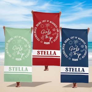 Könnte beinhalten: Drei Strandtücher in Grün, Rot und Marineblau, jedes mit dem Text "Girls Trip 2026" in einem kreisförmigen Design. Die Handtücher tragen auch den Namen "Stella" in weißen Blockbuchstaben.