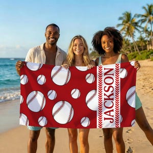 May include: A red beach towel featuring a pattern of white baseballs with red stitching. The towel has a white vertical stripe with the name "JACKSON" in red. The towel is held by three people on a beach.