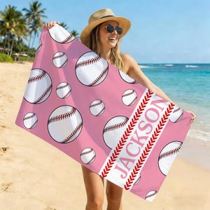 May include: Pink beach towel featuring a pattern of white baseballs with red stitching. The towel has a white stripe with the name "JACKSON" in pink letters and red baseball stitching. The towel is held up on a beach.