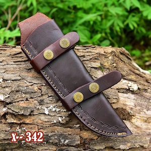 Può includere: Un fodero per coltello in pelle marrone con cuciture decorative e dettagli in ottone. Il fodero ha una patta con chiusura a scatto.