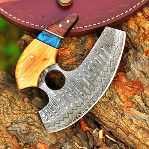 May include: A Damascus steel cleaver with a wooden handle and a blue and green epoxy resin accent. The cleaver has a hole in the blade and is resting on a piece of tree bark.