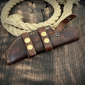 May include: A brown leather knife sheath with three brass snaps and a stitched seam. The sheath is on a wooden surface.