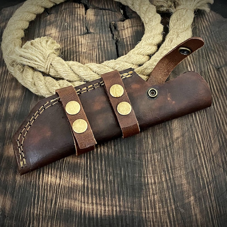 May include: A brown leather knife sheath with three brass snaps and a stitched seam. The sheath is on a wooden surface.