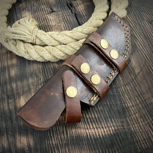May include: A brown leather knife sheath with a flap and snap closure. The sheath has brass rivets and stitching details.