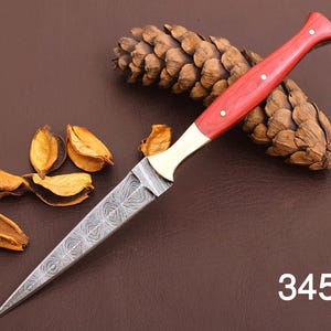 May include: A Damascus steel dagger with a red wooden handle and brass bolster. The blade has an intricate pattern. The handle is secured with three rivets. The number "3453" is visible in the lower right corner. The dagger is displayed with a pine cone and dried leaves.