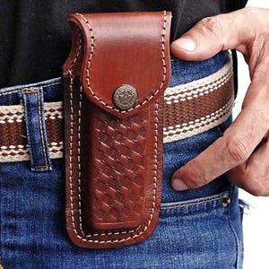 May include: A brown leather knife sheath with a basketweave pattern and a star-shaped button closure. The sheath is attached to a brown and white striped belt, worn with blue jeans. The stitching is white.