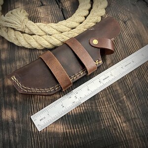 May include: A brown leather knife sheath with a stitched design and a brass button. The sheath is measured against a ruler.
