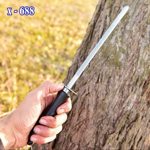 May include: A knife sharpening steel with a black handle and a silver sharpening rod. The text "X-688" is visible in the upper left corner. The tool is held in front of a tree trunk.