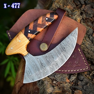 May include: A curved, stainless steel knife with a wooden handle. The handle is made of light wood with dark wood accents. The knife is in a brown leather sheath with a decorative design. The knife is labeled "X-477".