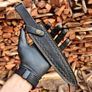 May include: A black leather knife sheath with a decorative scalloped pattern and gold stitching. The sheath has a strap with a brass button closure. The knife sheath is held by a person wearing a black glove.