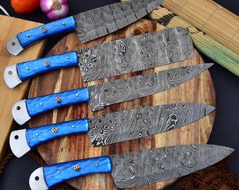 May include: Set of five blue handled kitchen knives with a Damascus steel blade pattern. The knives are arranged on a wooden cutting board.