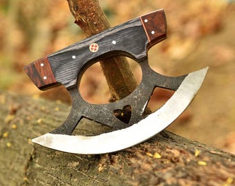 May include: A handcrafted, double-edged blade with a dark, textured handle and a heart-shaped cutout. The blade has a polished, curved edge. The handle features wood accents and decorative rivets. The knife rests on a tree branch.
