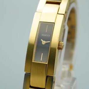 Puede incluir: Un reloj de pulsera Gucci dorado con una esfera rectangular negra. La esfera muestra el nombre de la marca y dos líneas doradas. El reloj tiene una correa dorada y una corona dorada.