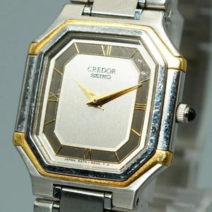 May include: A Credor Seiko wristwatch featuring an octagonal silver and gold-tone case with a light gray dial. The watch face displays gold-tone hands, Roman numeral markers at the twelve and six o'clock positions, and the text Credor Seiko.