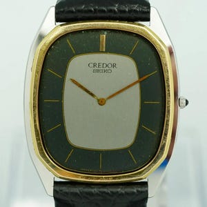 May include: A Credor Seiko wristwatch featuring a silver-toned center dial surrounded by a dark green border, gold-toned hands, and thin gold-toned hour markers. The watch has a gold-toned bezel, a silver-toned case, and a black leather strap.